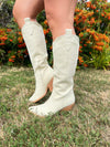 Beige cowboy boots worn outdoors on a grassy area with plants in the background