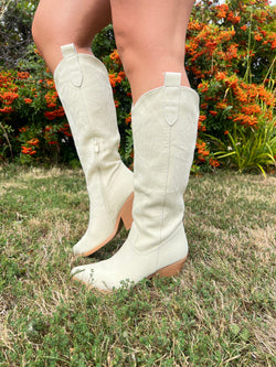Beige cowboy boots worn outdoors on a grassy area with plants in the background