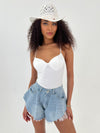 Woman wearing a white top and denim shorts with a white hat on a light gray background